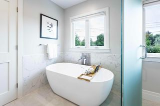 Freestanding white bathtub against a pale stone wall beside a window and glass shower enclosure.